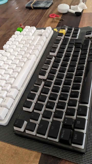 Dos teclados tkl iguales, uno blanco y uno negro, con un kit de limpieza