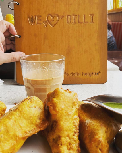 A plate of bread pakora in the foreground, a glass of masala chai in the middle, and a menu with I Heart Dilli carved on its wooden cover. A hand is holding up the menu behind the items.