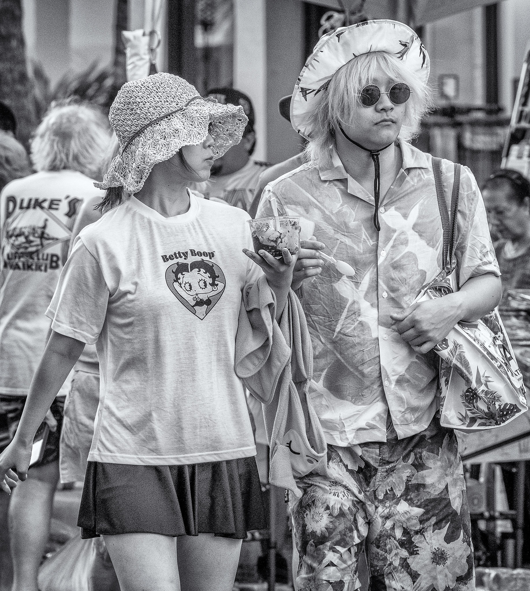 Couple walking along street near Waikiki in Honolulu. She has a Betty Boop t-shirt on ad is holding a frozen treat of some sort. He is dressed from head to foot in tropical-themed clothing, including hat and bag, wears round John Lennon style tinted glasses and is giving the photographer side-eye.