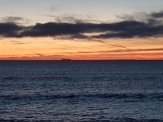 A large boat on the horizon, with pale orange glow behind it and a few darkened wispy clouds above.