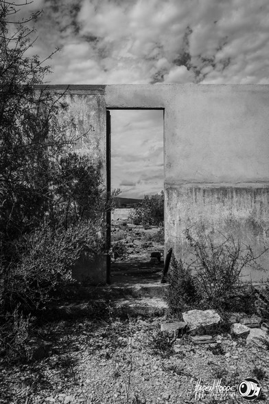 View through a door in a wall. The baraque is damaged and without a roof. Plants are in front of the building and behind the wall is the damaged concrete floor and the landscape in the background.