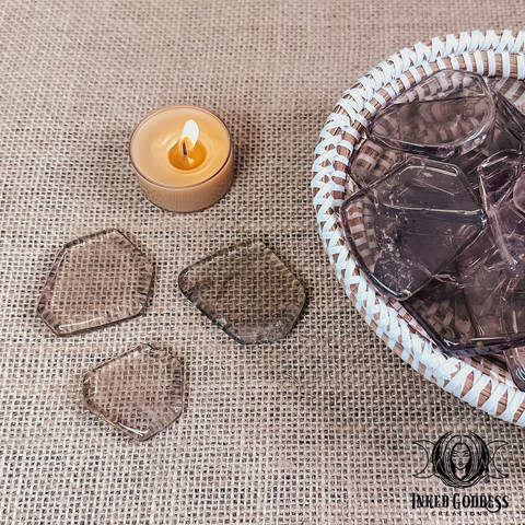 A photo of our "Smoky Quartz Slice" product. It features several specimens in a wicker bowl with three specimens on the burlap-covered surface next to the bowl. A gold tealight candle is shown for scale. Presented by Inked Goddess Creations.