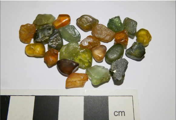 A cluster of 25 small sparkly, transparent, gem-like pebbles of zircon, in various shades of green, brown and yellow, on a white paper background, with a black-and-white cm scalebar at the bottom  of the picture.