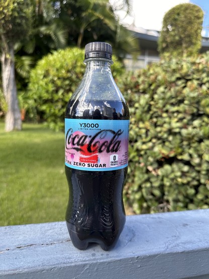 A bottle of Coca-Cola Y3000 outside on a fence in front of a garden. 