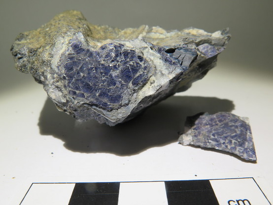 Two pieces of rock sit on a white paper background, starkly illuminated in raking light which creates strong shadows. On the brightly illuminated pafes of the rocks, platy crystals of blue sapphire can be seen. A black and white cm scale bar is visible at the bottom of the picture.