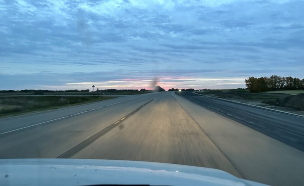 Foto aus dem fahrenden Auto. Grauer Morgenhimmel, aber am Horizont zeigt sich hell rötlich die demnächst aufgehende Sonne. In der Mitte des Bildes hat es ein wenig Flecken von verschmierten Insekten auf der Windschutzscheibe.