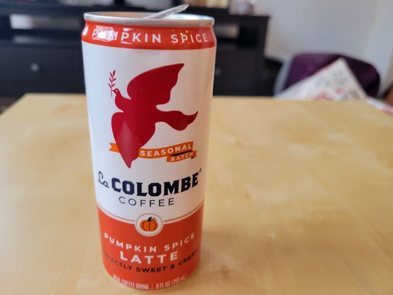 9 fluid ounce can of La Colombe Pumpkin Spice Latte Coffee, on a birch veneer IKEA LACK side table. The La Colombe can has a white background with orange borders at top & bottom, and a dark orange silhouette of a dove of peace in flight while holding an olive branch, flying above a banner reading "Seasonal Batch". Below this is a small pumpkin enclosed in a gold circle with a white background. Other text on the can: "Perfectly Sweet & Creamy".