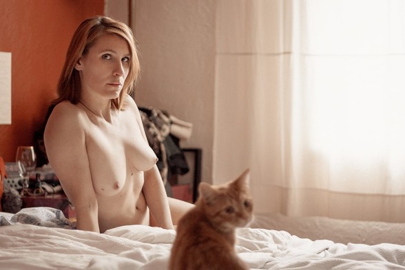 Nude woman sitting on her bed next to her cat, both looking in the camera's direction