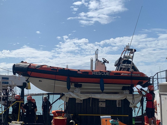 Das RHIB steht auf einer Tragekonstruktion, Menschen drum herum. Das RHIB sieht wie neu aus. Weißer Rumpf, orangene Schläuche. Das Wort RESCUE ist zu lesen.