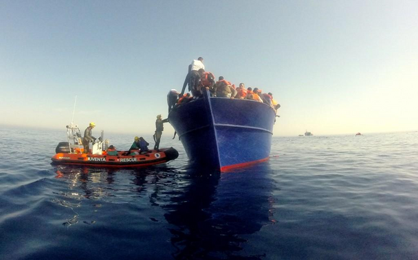 Das RHIB iuventa-rescue bei einem hoch aufragendem einfachen Holzboot, das voller Menschen ist und zum RHIB hin krängt.