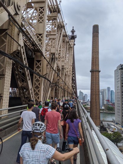 A large group of people, many with bicycles, walks over the southbound outer roadway of the queensboro bridge, a large steel cantilever bridge. In the distance we can see the bridge passing over the river, next to a large brick smokestack that is taller than the bridge itself.
