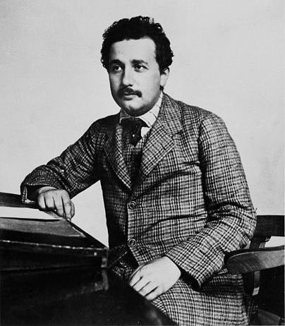 Einstein at age 25, in a tweed suit. He is seated at a desk with his right arm resting on a podium or lectern. He has dark hair, and is looking to the left of the photographer in this posed photo.
