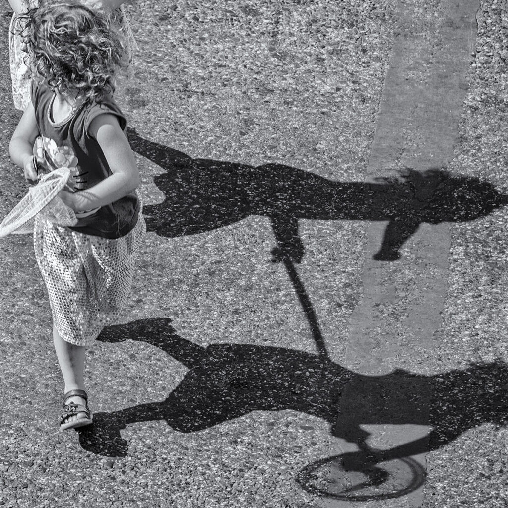 Two girls tussle over a butterfly net.Their shadows project on the roadway as they run across the street.
