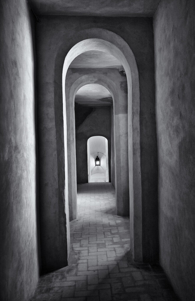 A arched hallway with a lamp visible at the end. Shadows from windows trace a serpentine pattern on the tile floor.