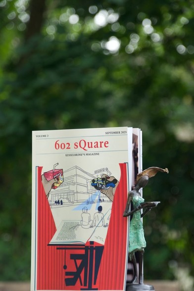 a picture of the magazine's front cover with a drawing of people sitting at Kesselbrink on a big magazine's page, two hands entering the cover holding a Durstlöscher and smashed police car.
Besides the magazine there's a small statue holding something, leaning against the pages.
Blurry trees in the background.