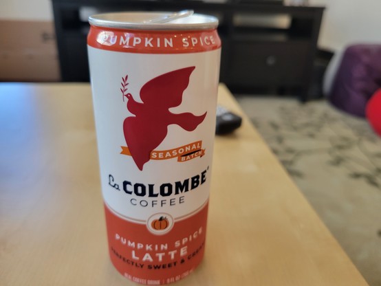9 fluid ounce can of La Colombe Pumpkin Spice Latte Coffee, on a birch veneer IKEA LACK side table. The La Colombe can has a white background with orange borders at top & bottom, and a dark orange silhouette of a dove of peace in flight while holding an olive branch, flying above a banner reading "Seasonal Batch". Below this is a small pumpkin enclosed in a gold circle with a white background. Other text on the can: "Perfectly Sweet & Creamy".
