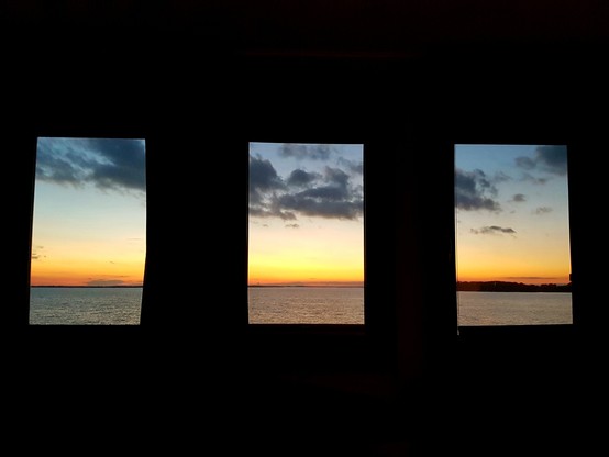 Blick durch ein schwarzabgedunkeltes Zimmer auf drei Fenster, die den Blick auf einen Seehorizont mit orangerotem Abendhimmel und dunkelblauen Wolkenfetzen freigeben.