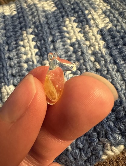 Tiny unidentifiable creature made out of borosilicate glass held between fingers. It has two ears, a long-ish nose or face, and a weird bowling-pin looking body. It’s sort of yellow but mostly clear. 