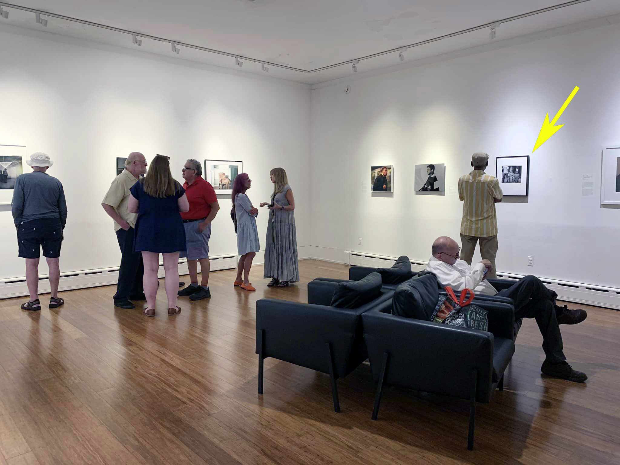 Image of museum reception for photography exhibition. Exhibition photos are arranged on the walls and clumps of people stand before them, alternatively talking amongst themselves and admiring the photos.