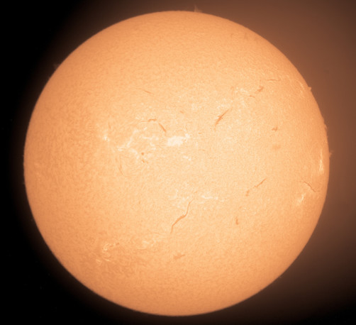 Round yellow-orange disk of the sun fills the image view.  There are a couple small flame- like prominences coming off the edge.  On the surface there are some dark lines of filaments, white active areas of plage and some dark spots.