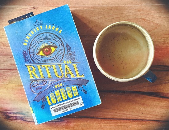 Holztisch von oben mit Buch „Das Ritual von London“ von Benedict Jacks und Kaffeetasse. 