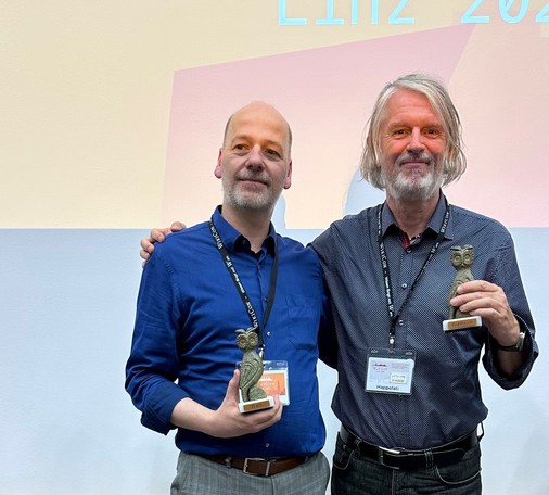 Zwei Wikipedianer, beide über 50, halten ihre Ehren-Eulen in den Handen. Der eine, ältere legt seinen Arm um den jüngeren. Der Gesichtsausdruck von beiden drückt ihre Freude über den verliehenen Preis aus. 