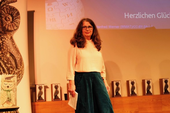 Die Laudatorin von zwei Preisen steht auf der Bühne und sieht ins Publikum. Sie hat lockige Haare bis über die Schulter, eine Brille und eine zarte rosa Chiffonbluse zu einer blaugrünen Cordhose an. 
Foto: Kathrin Herwig
