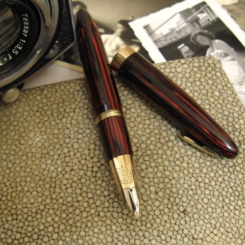 An uncapped vintage Sheaffer fountain pen, cap by its side, surrounded by vintage ephemera (a cigarette case, old photographs, and a Rolleiflex camera).