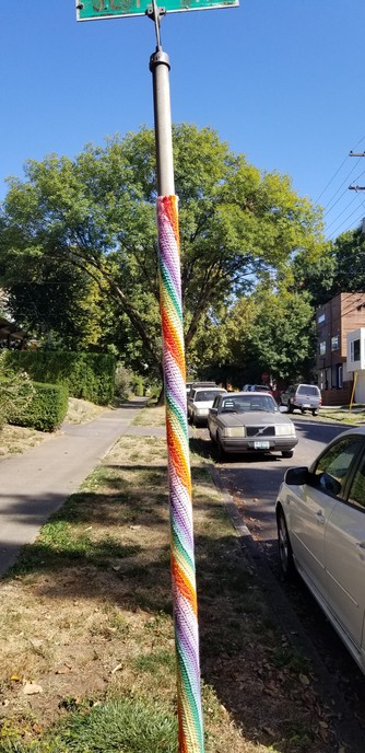 A street sign has been yarn bombed with rainbow in a spiral up the pole.