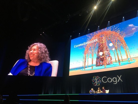 A photo of the CogX Festival showing on stage 3 women small in comparison to the giant projector screens behind them showcasing on the left screen Diana Ayton-Shenker, CEO of Leonardo/ISAST, a light-skinned woman with peppered shoulder-length hair and a radiating smile on her face as she looks out to the audience. She is wearing a black shirt with royal blue blazer overtop and a large drooping gold necklace. On the large screen on the right is a photo of the XR Art by William T. Ayton called ConstellationXR. Below this screen is the festival logo CogX. Below this logo are the 3 women on stage, barely visible to the eye.
