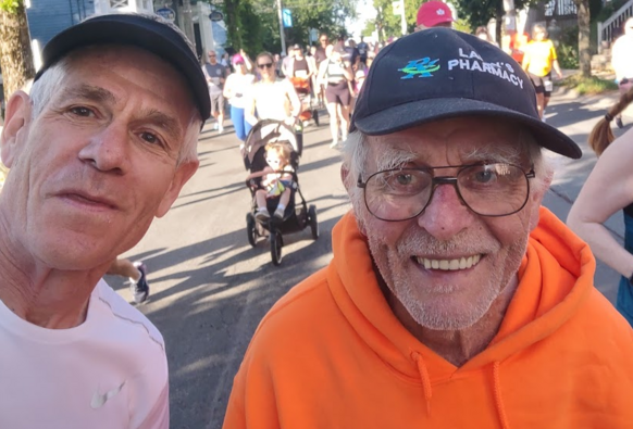 Selfie of me wearing a white jersey and another gentleman who is wearing a bright orange sweatshirt, glasses and a baseball cap. It is near the start of a road race. Other runners, including a woman pushing a stroller, can be seen in the background.