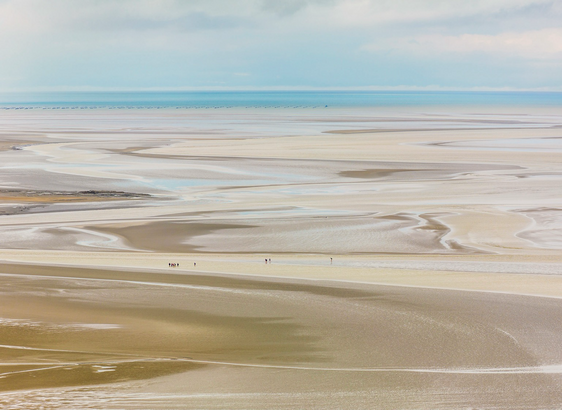 Paesaggio di una spiaggia bretone durante la marea bassa con persone in lontananza che stanno camminando tra le pozzanghere e il fiume. All’orizzonte il mare con le coltivazioni di ostriche. Il tutto in toni pastello.