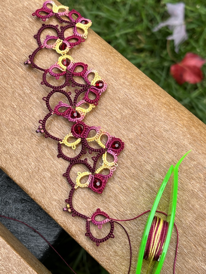 Beaded tatting on bench outside in red, pink, and yellow thread.