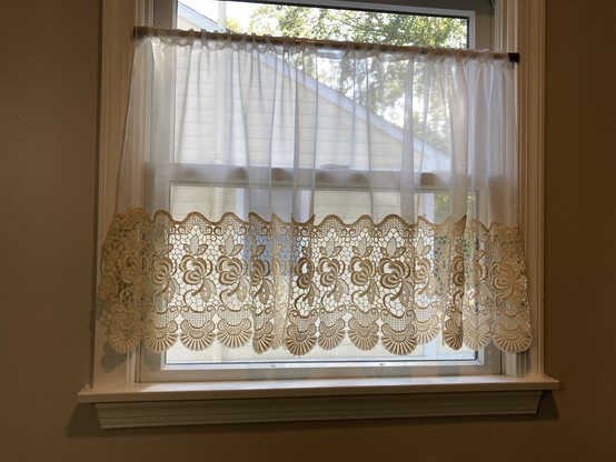 Translucent white curtain with cream lace along the bottom, hanging from a tension rod in a bathroom window 