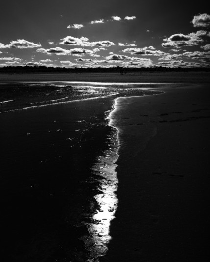 Black and white photo of the water’s edge on a beach. The very edge of the water where it meets the sand is illuminated by the sun (which is out of frame) and takes the form of an irregular  vertical path that leads the viewers eye from the bottom/foreground of the image up towards the horizon, and then  curves first right then left below the upper part of the beach. Above the beach, the sky displays a clutch of fair weather clouds, each one rim lit by the sun behind. The whole image is rendered with a very constrasty tone, with the sand, water and sky very dark and the reflections on the water and edges of the clouds very light.