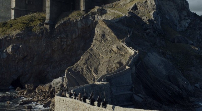 A rocky island with a dramatic and winding stone staircase. The image is from the Game of Thrones television show. The show producers have changed the look of the island to make it larger and more looming, with a large computer graphic castle on top that looks menacing.
