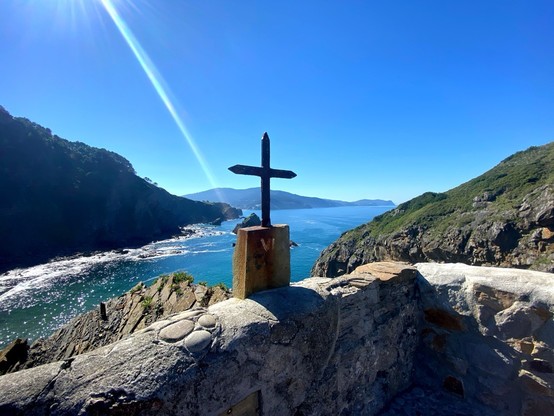 From atop a stone platform, a vast expanse of land and sea lays out before us. To the right, the rocky, grassy outcropping of the island where we stand. To the left, the looming, dark bulk of the mainland. In the middle, the blue-green sea with waves washing on the shore. Immediately in front of us, the low stone wall that keeps us from toppling over the edge. On that wall, in the middle of our view, a simple iron cross sits on a wooden base. From the sky, the sun sends out a single ray of light, created by lens flare.