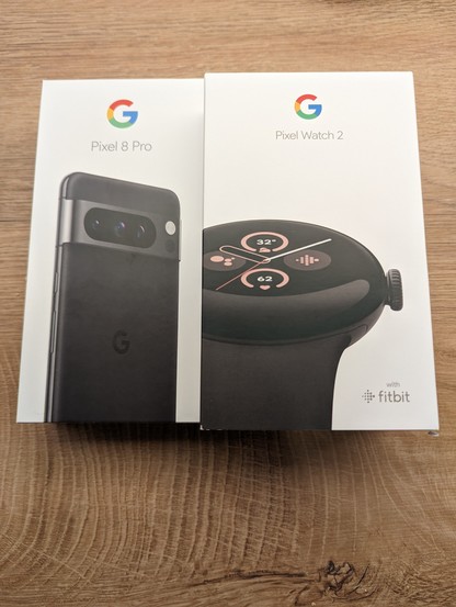 Bild der verschlossenen Originalverpackung einer Google Pixel Watch 2 rechts im Bild und links im Bild vom Google Pixel 8 Pro 