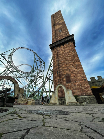 Photo of Kärnan roller coaster at Hansa-Park. Taken from ground level to emphasise the height of the green roller coaster track, mess of green supports, and very tall brick tower.