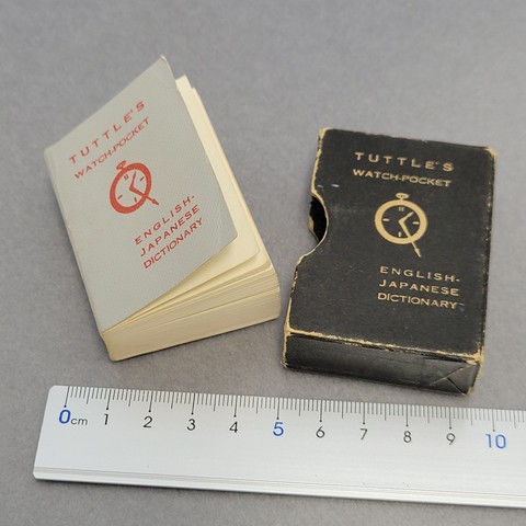 Tiny watch-pocket English-Japanese dictionary lying next to its cover box and a ruler for size comparison.