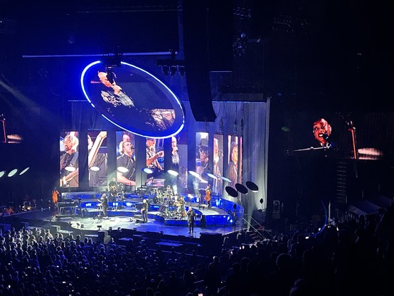Audience shot from a Peter Gabriel concert. A sea of heads in the foreground, the stage and big screens behind. 