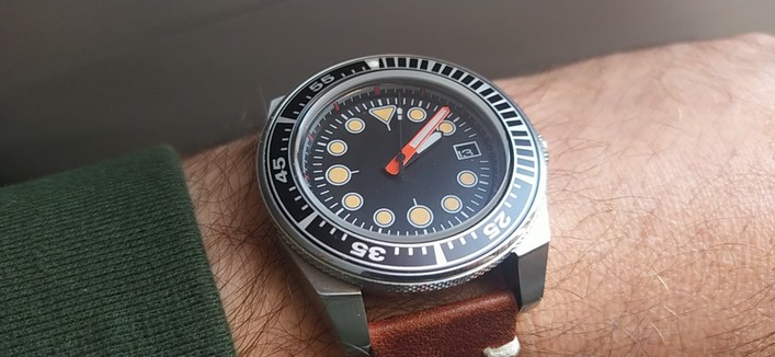 A bezelled dive watch with a brown leather strap. The black dial is ornamented with brass dots. 