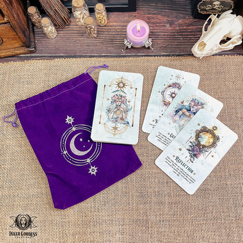 A photo of our exclusive "Mystical Messages Oracle Deck" product. It features three cards laid out on a burlap-covered surface. The rest of the deck is in a single stack atop a velvet drawstring bag with moon and star imagery. A purple tealight candle is shown for scale. Presented by Inked Goddess Creations.