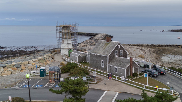 Scituate Lighthouse in the foreground, covered in scaffolding, and with the beacon housing set on the ground.
