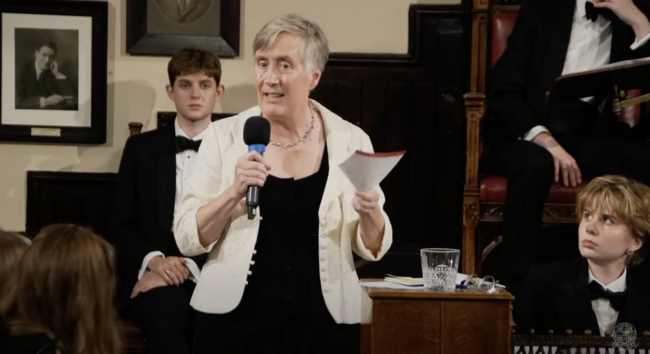 picture of me in a black dress, necklace and white jacket holding a paper and a microphone, with two students in tuxedos (one male, one female) looking at me, and a  mid 20C black & white picture of some white guy posing on the back wall. Actually, we are all white in the picture, but there was decent ethnic diversity in the audience.
