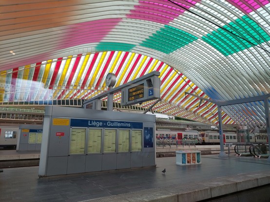 Blick aus dem Zug auf den Bahnsteig in Liege - Guillemins. Im Vordergrund sieht man eine Wand, auf der die Zugverbindungen zu sehen sind und darüber Hinweistafeln und die Bahnhofsuhr. Besonders ist an diesem Bahnhof aber die Dachkonstruktion, die halbtransparent in vielen Farben leuchtet.