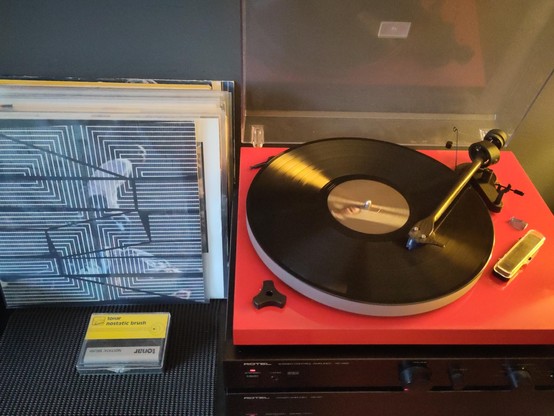 A bunch of records leaning against a grey wall. The front record sleeve, front cover showing, is of David Grubb's record 'The plain where the palace stood'. To the right, a stereo set consisting of a black power amplifier (bottom), a black stereo control amplifier (middle) and a bright red record player (top), playing the aforementioned record, B side.