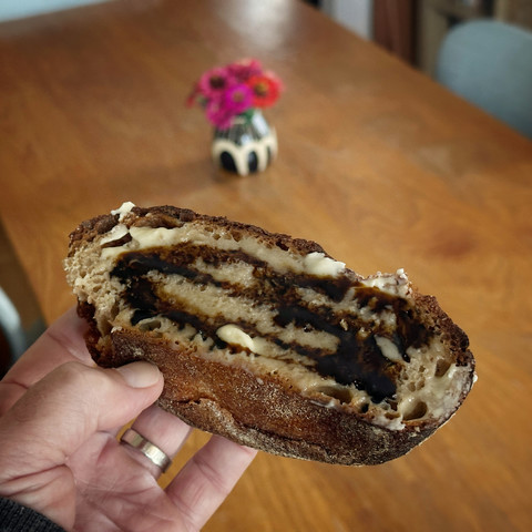 Photo of a heel of sourdough bread, held in a hand. The bread is butter and molasses on it. There is a table in the background with a small bouquet of flowers on it.