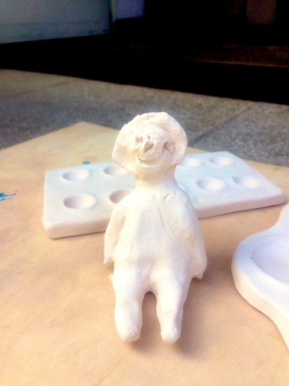 Auf einem Holzbrett liegt eine getöpferte Mischpalette. Davor sitzt eine kleine Figur, die wie ein Baby aussieht und von einem 4-jährigenMädchen geformt wurde. Beides ist weiß und noch nicht glasiert.