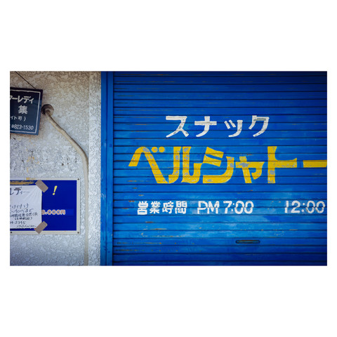 The image depicts a vibrantly blue shutter of an establishment in Nagoya. Bold yellow characters on the shutter signify it as a "snack" bar. Adjacent to the shutter, there's a cracked, white wall with a sinuous air conditioner conduit and two faded notices attached. Despite its name, a "snack" bar in Japan typically refers to an entertainment venue, often for middle-aged men attended by young female hostesses, rather than a place to get bite-sized food items.